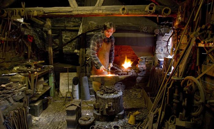 Inside the blacksmith's workshop. There are lots of tools strewn around and a forge burning brightly in the background. The blacksmith wears a leather